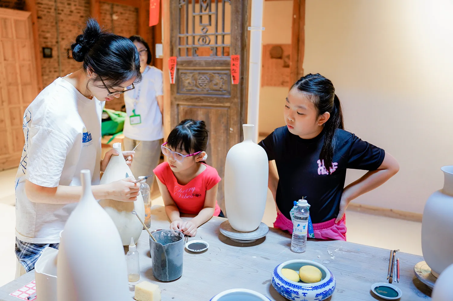 Two girls watch an instructor paint an unfired porcelain vase with a fine brush in a Jingdezhen studio