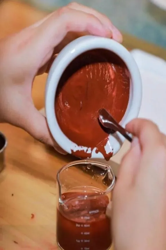 Red pigment slurry poured from a ceramic bowl into a small glass beaker