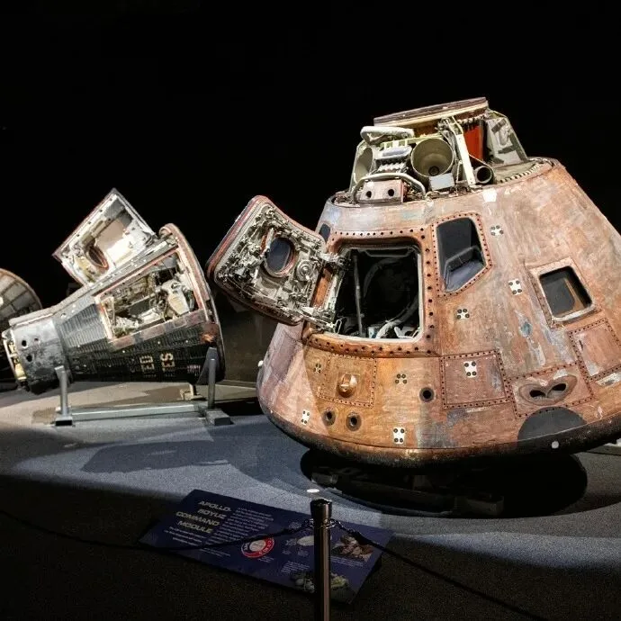 A scorched Apollo command module and a second capsule on display in a darkened museum hall.