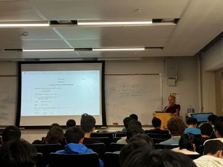 Students in a tiered lecture hall facing a projected course syllabus and an instructor at the podium.