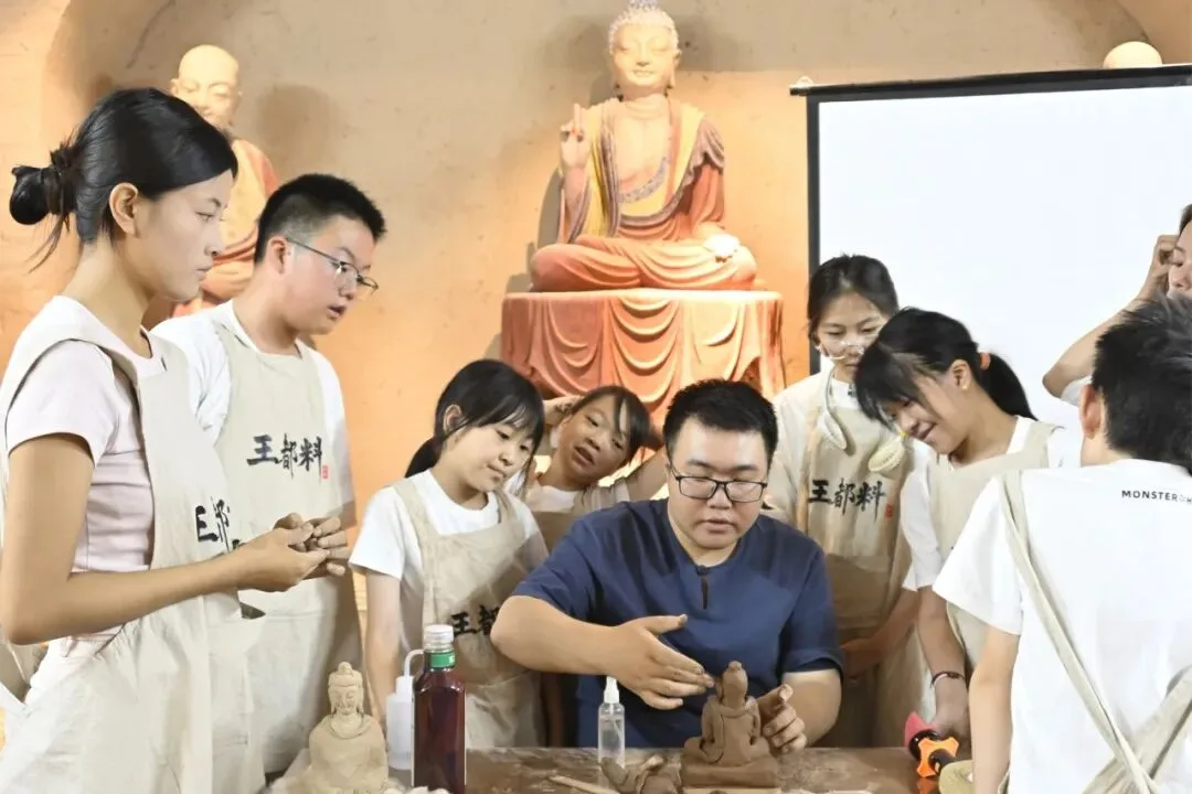 Instructor shaping a small clay figure as children watch, a seated Buddha statue behind