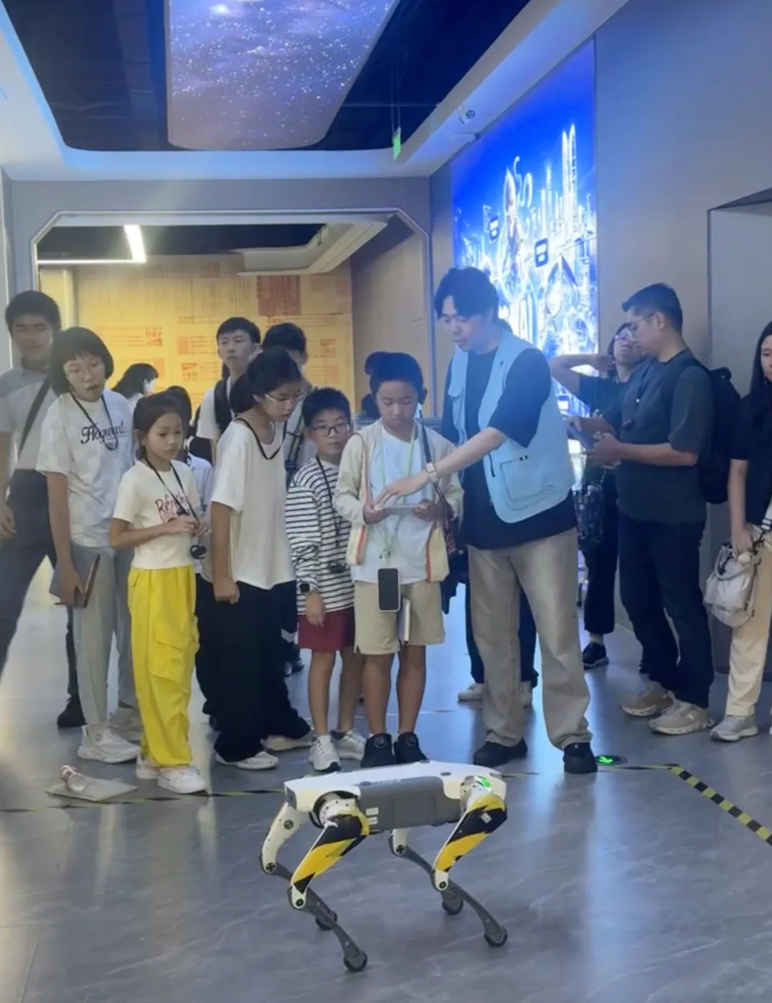 A guide demonstrates a yellow-and-white quadruped robot walking in front of the children