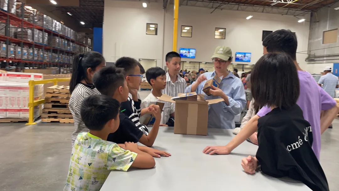 Students gathered around a table in a food-bank warehouse as a volunteer leader demonstrates packing.