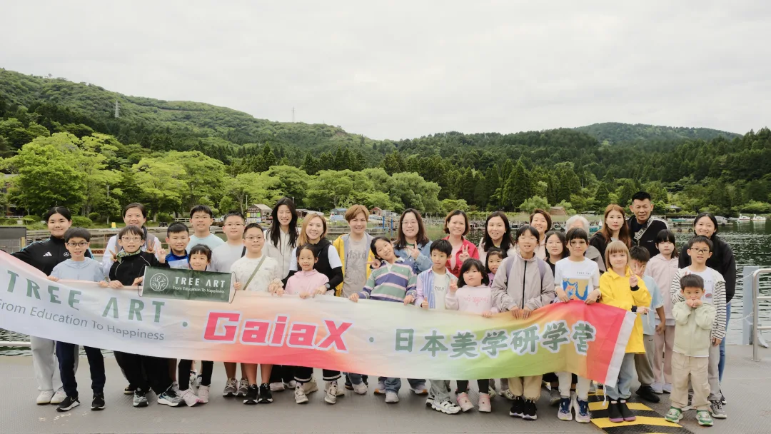 Students and parents by a lake holding a TREE ART and GaiaX Japan aesthetics camp banner