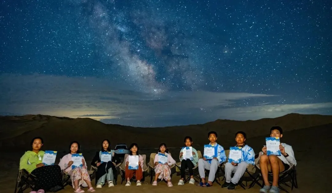 Nine children seated in camp chairs on the desert holding certificates under the Milky Way