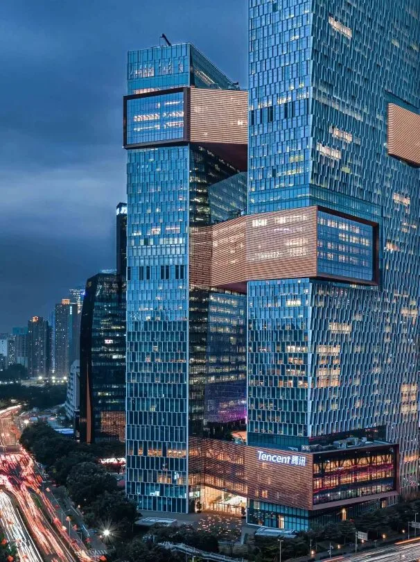 Tencent's twin Binhai towers linked by bridge blocks, glowing over Shenzhen traffic at dusk.