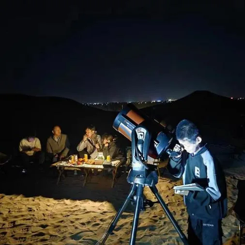 A boy at a large telescope on the night dunes, others seated at a lantern-lit table behind