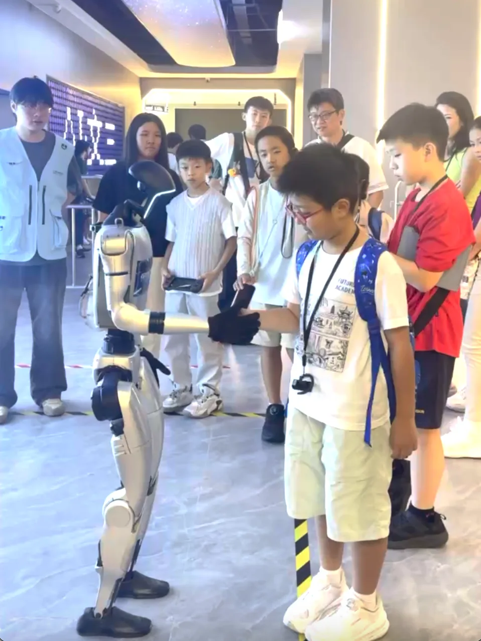 A boy shakes hands with a white humanoid robot as his classmates watch