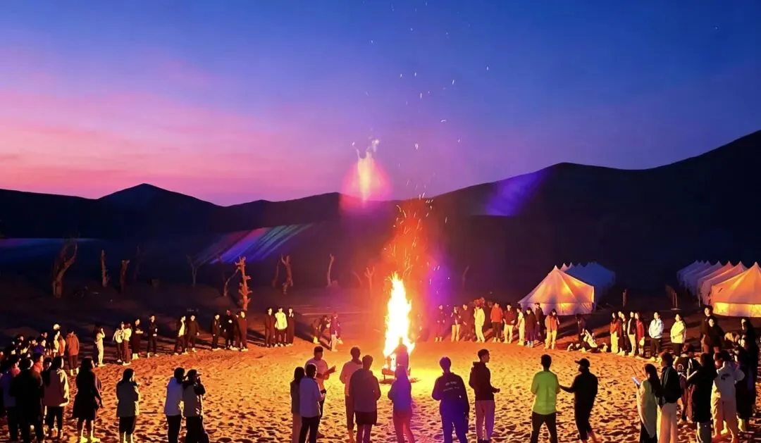 Crowd ringing a tall bonfire on the sand at dusk, white tents to one side