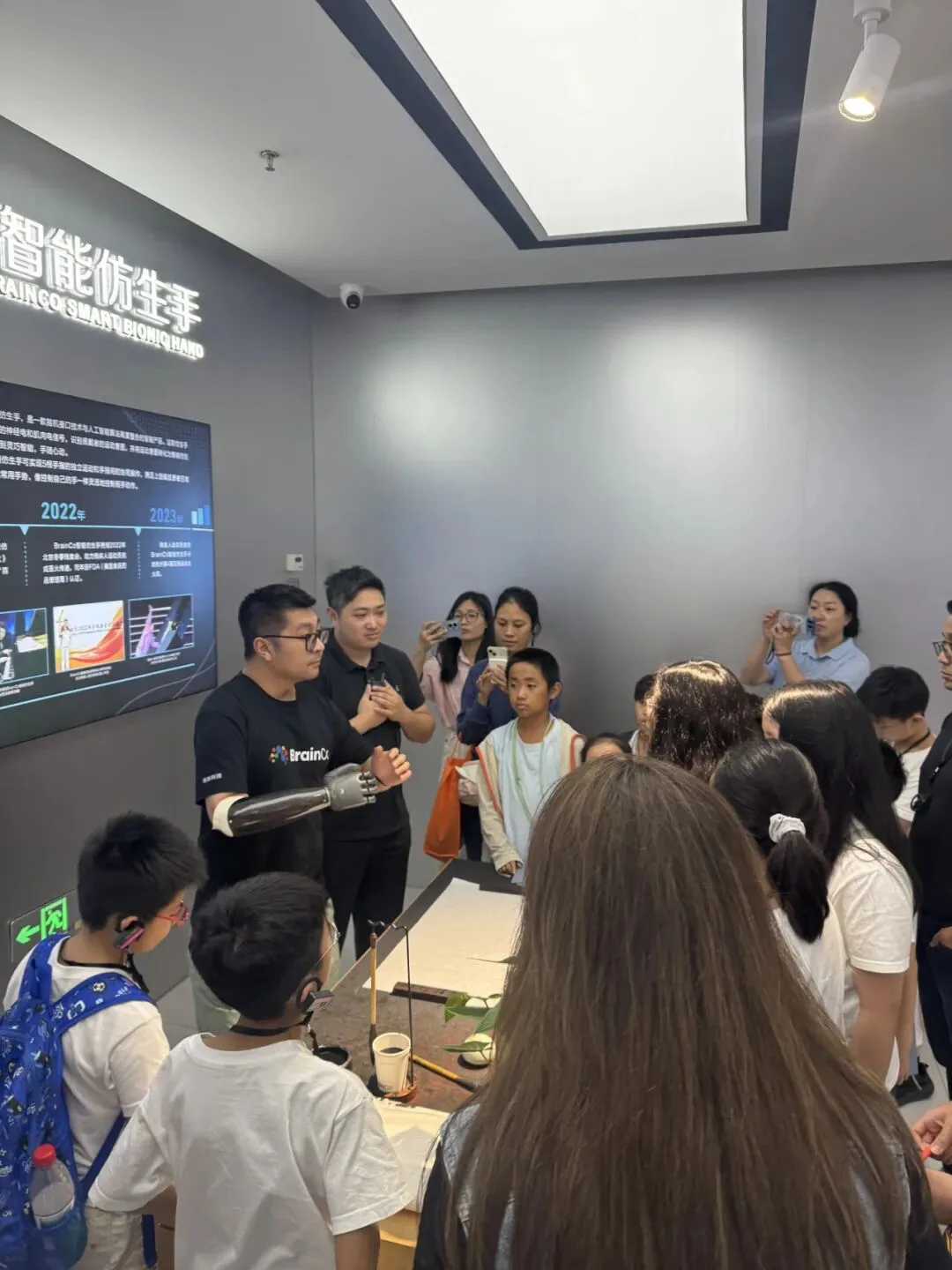 A presenter wearing a smart bionic hand demonstrates it to children beside its display wall