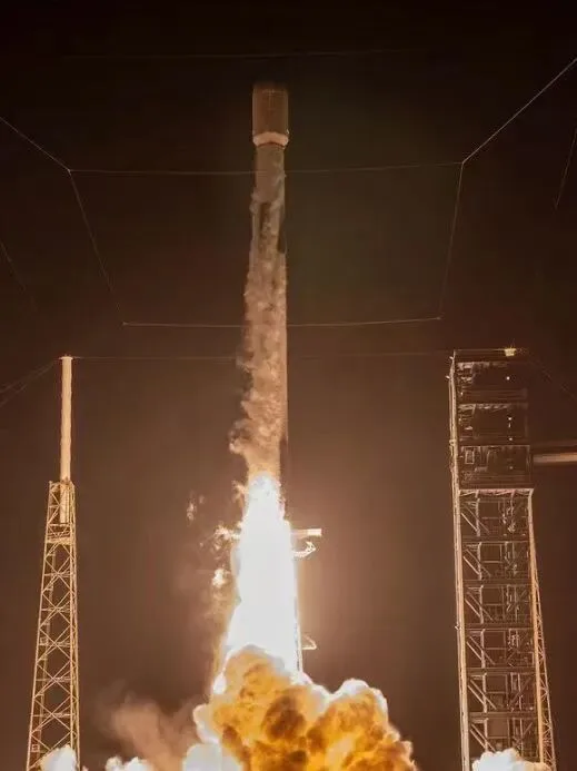 A rocket lifting off at night in a plume of flame between launch-pad towers.