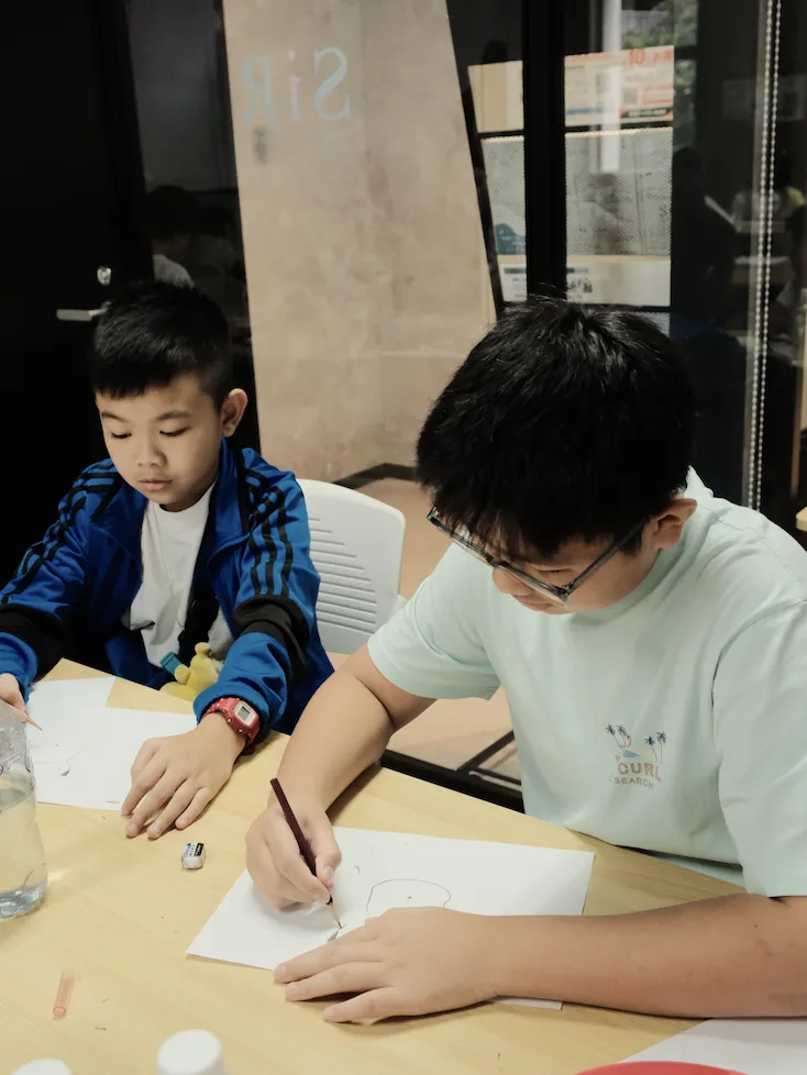 Two boys drawing character outlines in pencil at a classroom table