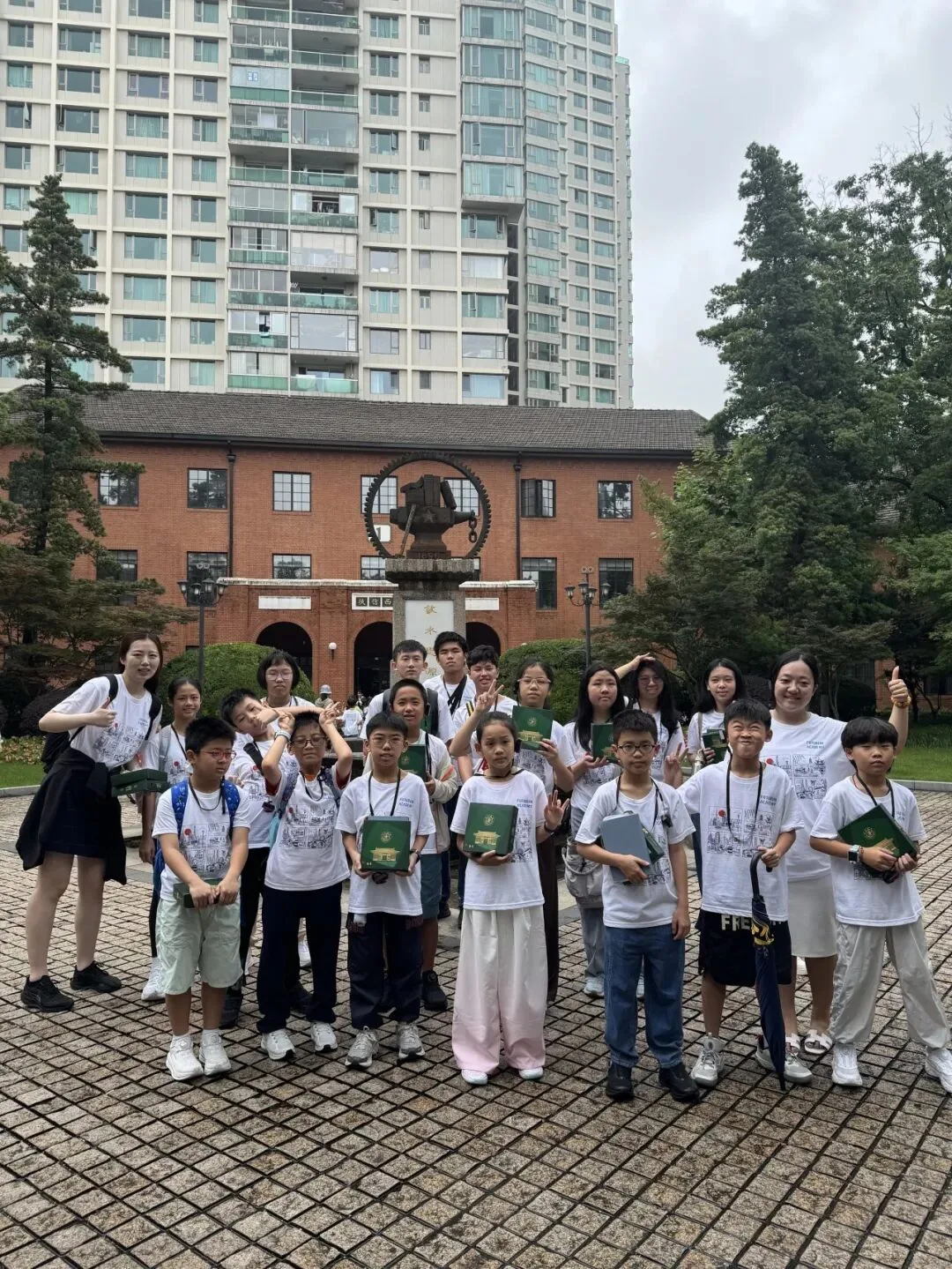 The group poses with green notebooks in a red-brick campus courtyard beneath a bronze monument