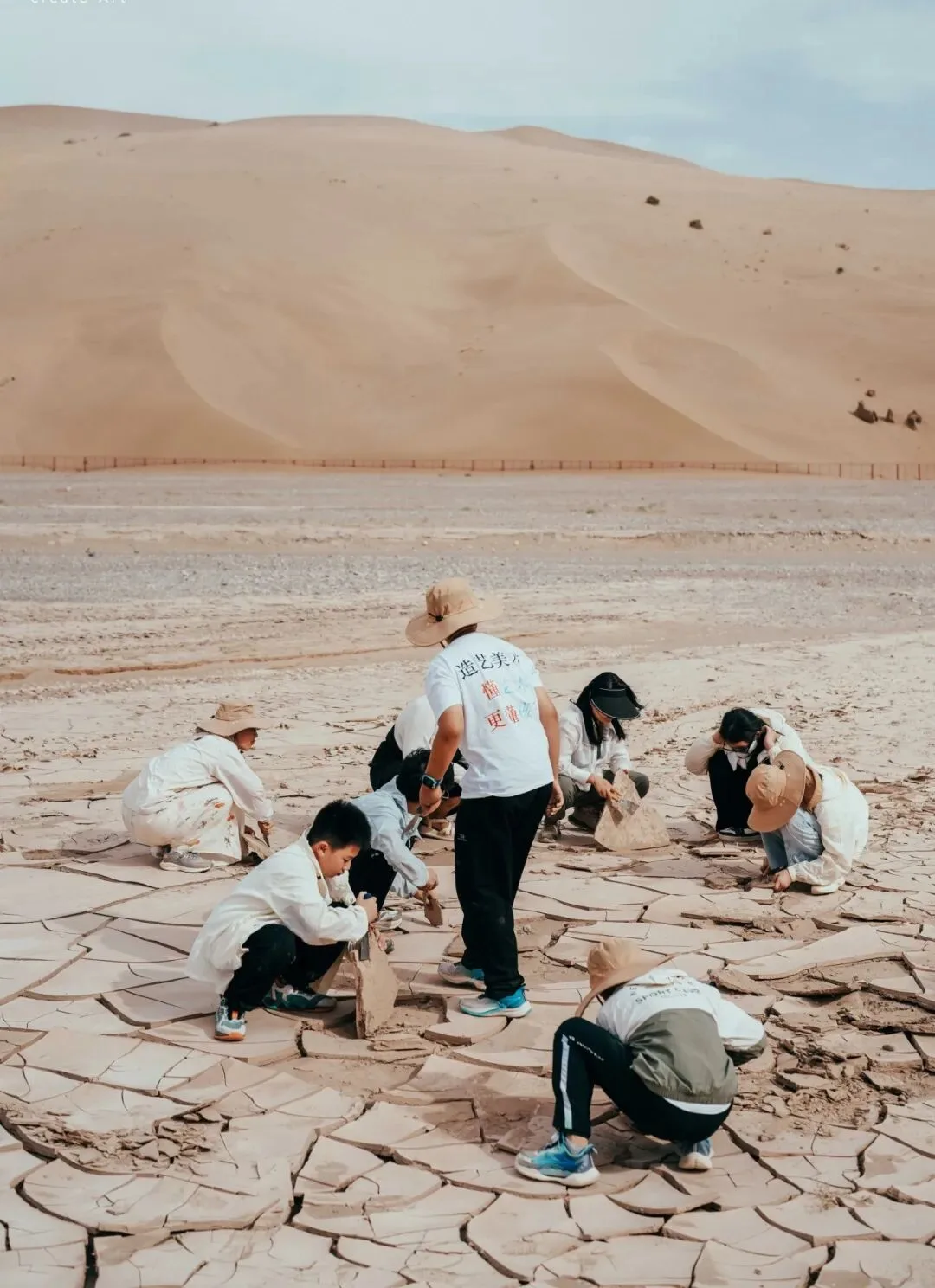 Group crouched on cracked clay flats prying up slabs, a tall dune behind them