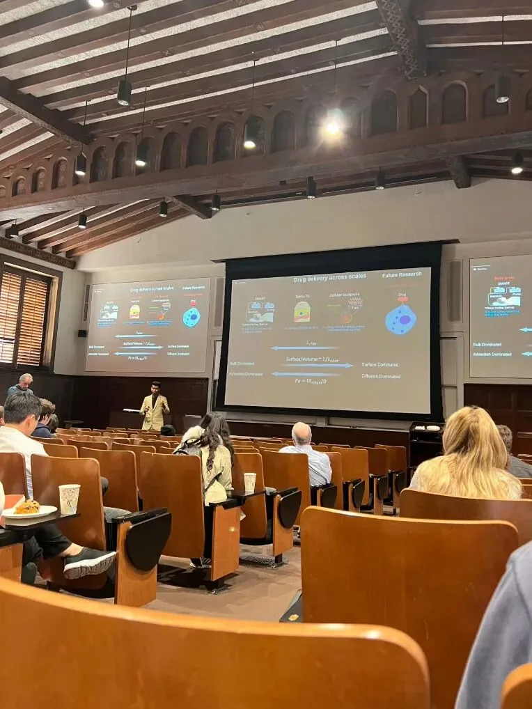 A presenter beside screens reading Drug delivery across scales in a wood-beamed lecture theatre.