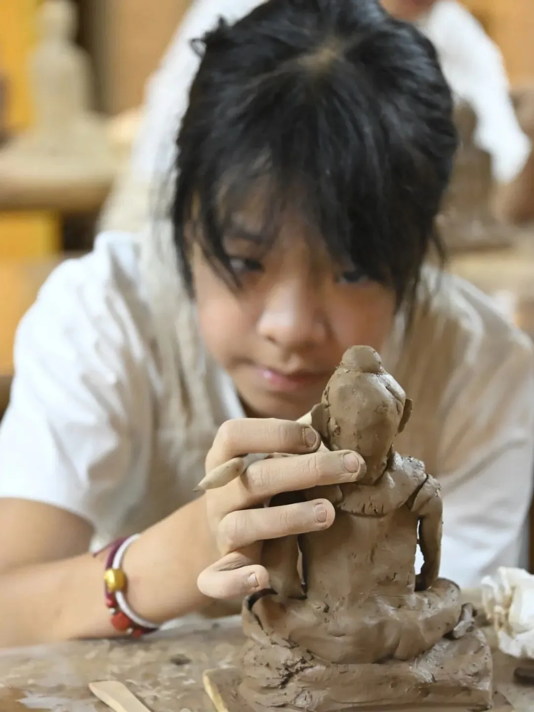 Child shaping a small seated clay figure with fingers and a wooden tool