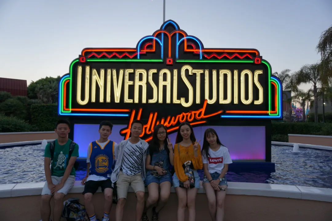 Six students sitting in front of the neon Universal Studios Hollywood sign at dusk.