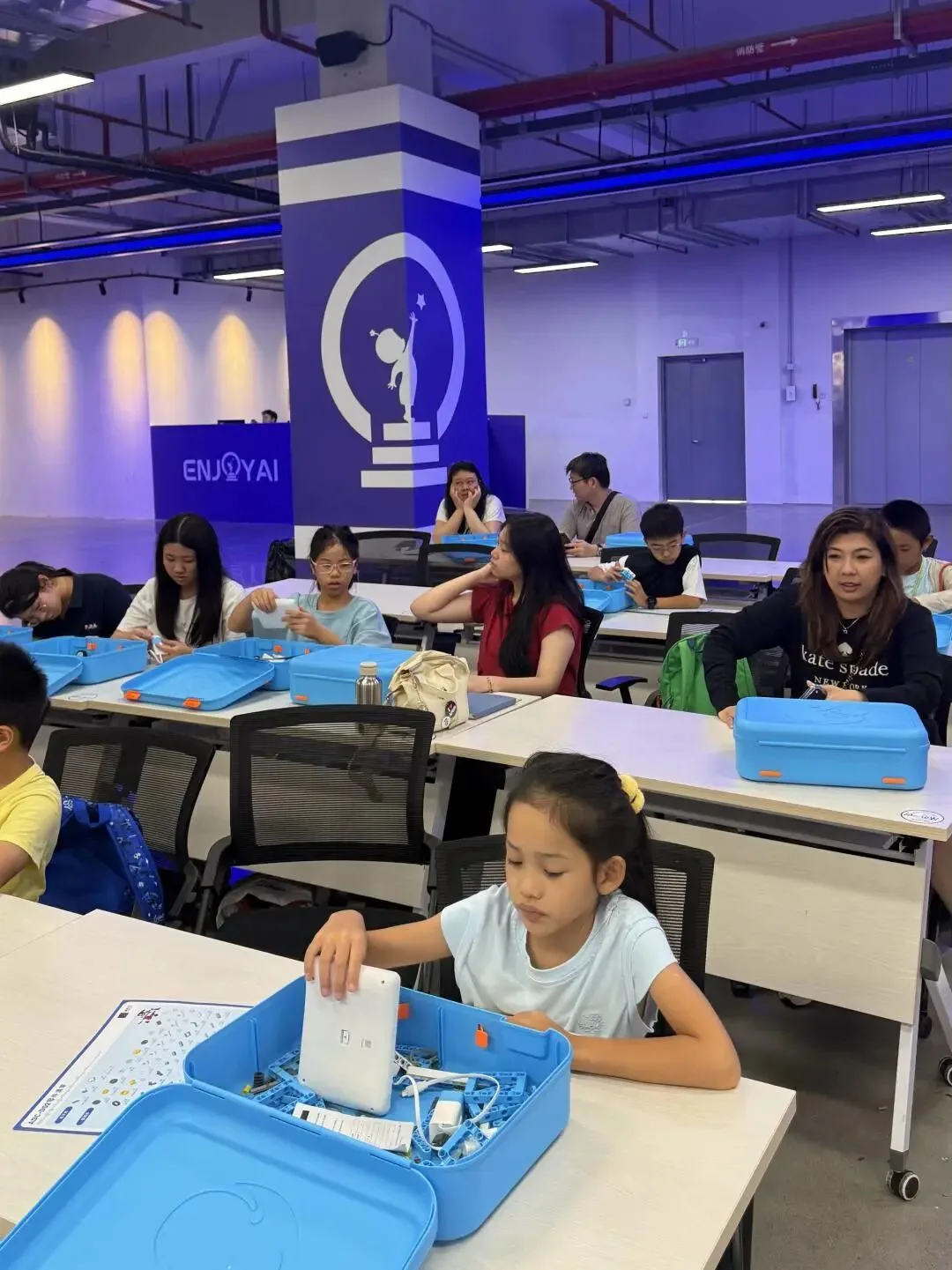 Children at desks open blue robot kit boxes in the EnjoyAI training classroom
