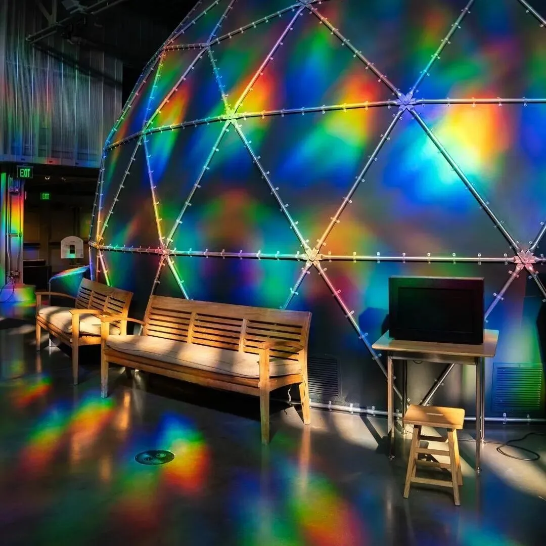 Benches and floor washed in rainbow light beneath a lit geodesic dome.