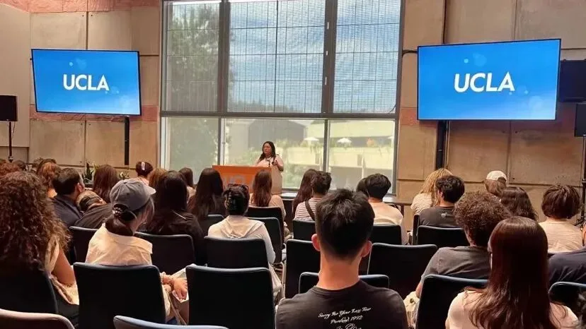 An audience seated in a UCLA hall watching a speaker at a podium between two UCLA screens.