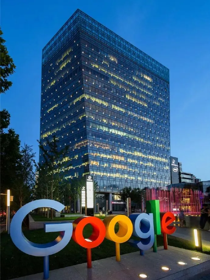 A multicoloured Google sign in front of an illuminated glass office building at dusk.