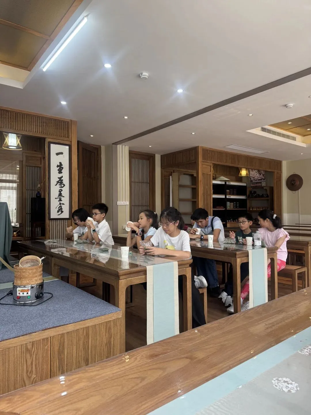 Children taste tea at long wooden tables in a tea house hung with a calligraphy scroll