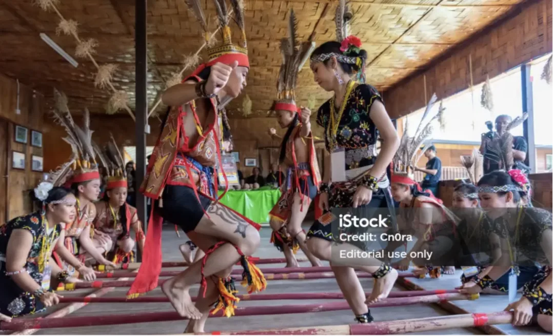 Dancers in feathered headdresses and beaded costume step between clapping bamboo poles inside a longhouse.