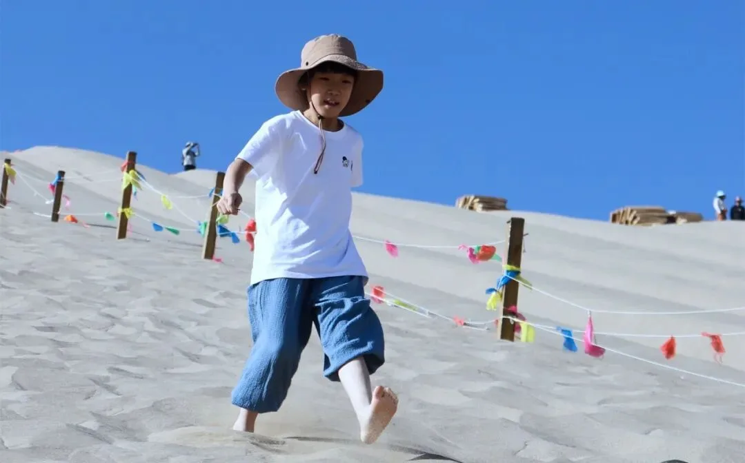 Barefoot boy in a sun hat running down a pale sand dune past coloured bunting
