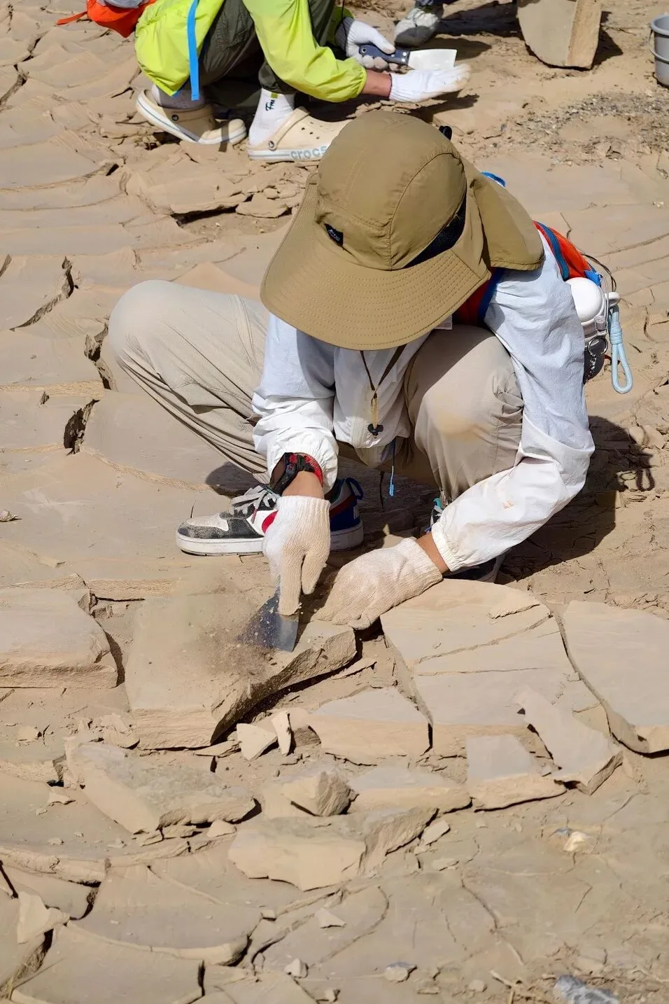 Child in a sun hat and gloves prying pale clay slabs from cracked desert ground with a trowel