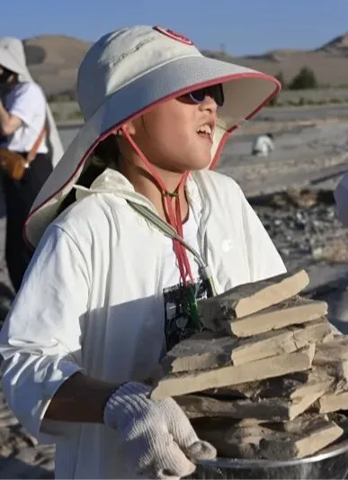 Girl in a wide sun hat and work gloves carrying a stack of pale clay slabs in a basin