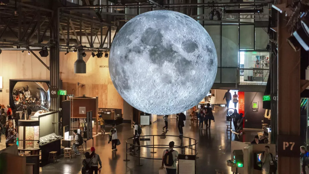 A giant illuminated moon sculpture suspended above visitors in a science museum hall.