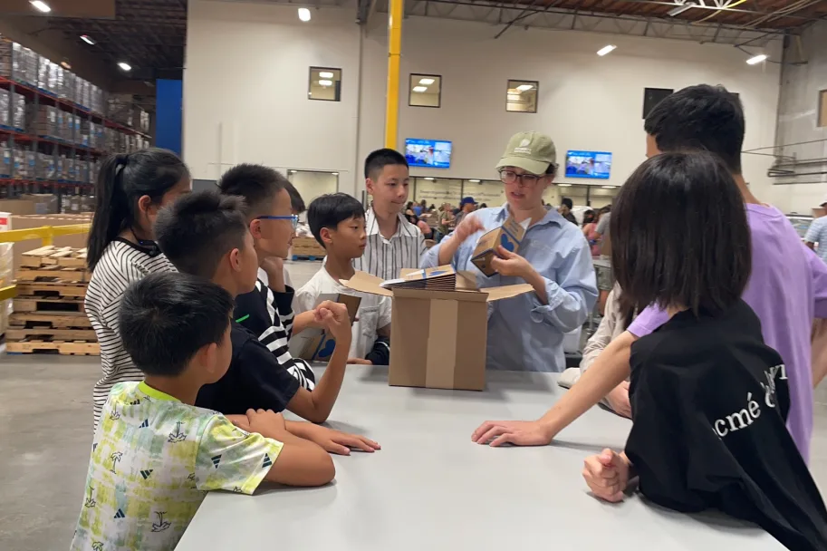 A volunteer leader showing a packing box to students around a table in a donation warehouse.