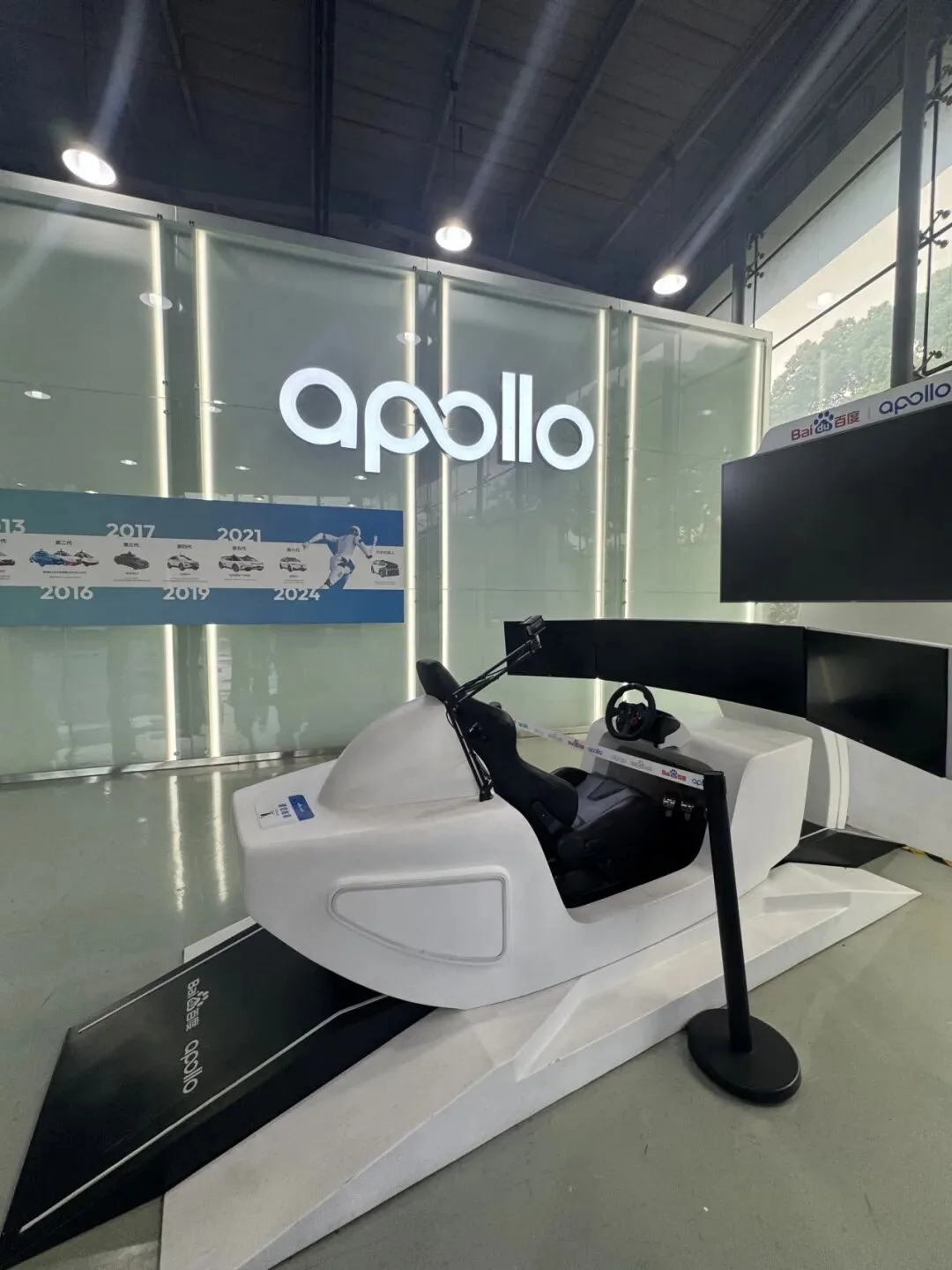 A white driving simulator pod sits beneath the Apollo sign beside a 2013 to 2024 timeline