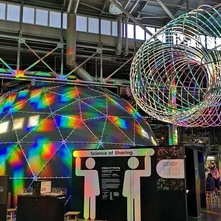 A geodesic dome washed in rainbow light beside a Science of Sharing exhibit sign.