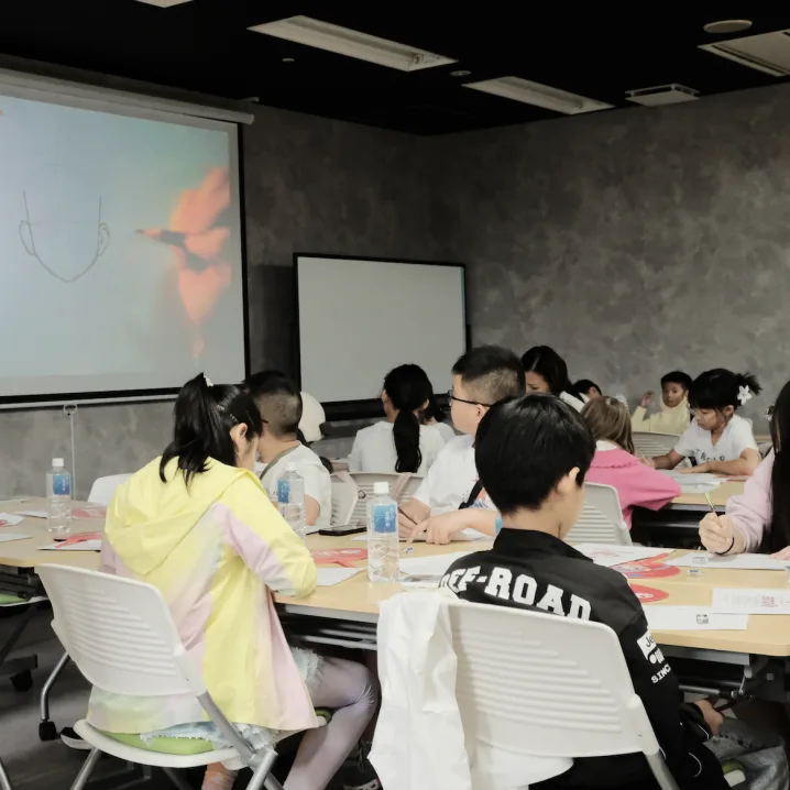 Children sketching at long tables while a drawing demonstration plays on the projector screen