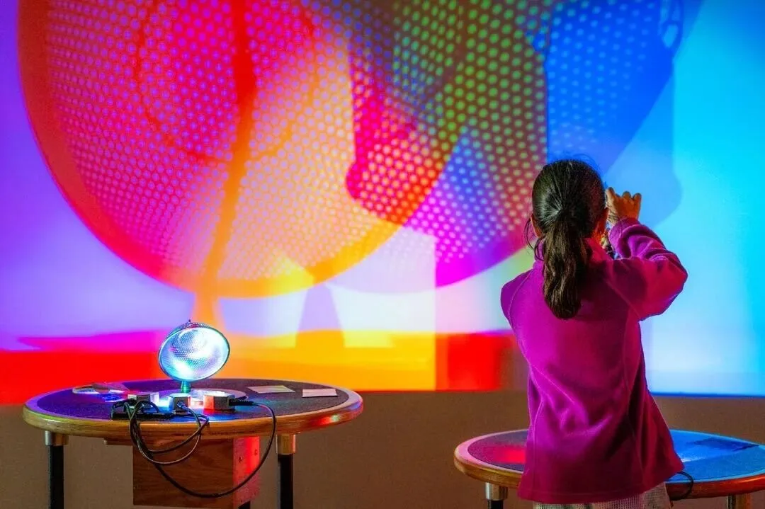 A child reaching towards overlapping bands of coloured light at a colour-mixing exhibit.