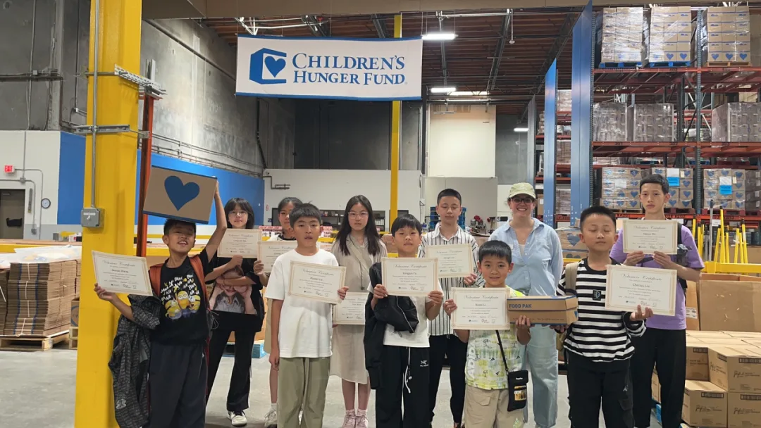 Students holding volunteer certificates beneath a Children's Hunger Fund banner in a warehouse.