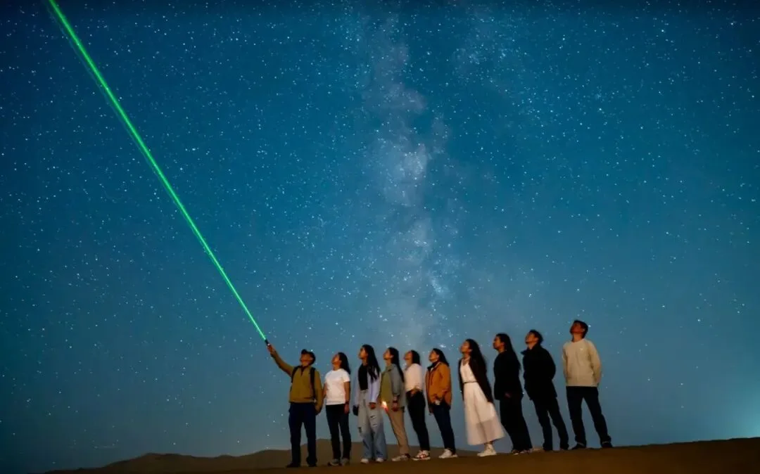 Group lined up on a dune under the Milky Way, one pointing a green laser at the sky
