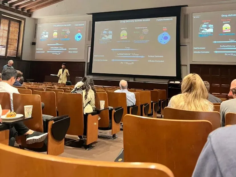 A wood-beamed lecture theatre with three screens showing a Drug delivery across scales talk.