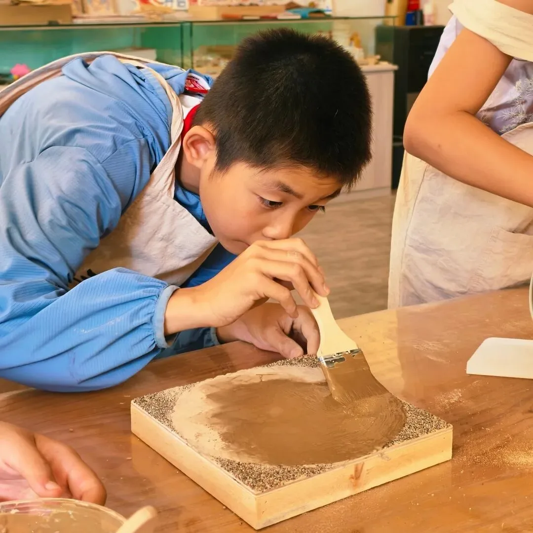 Boy in a blue smock brushing wet clay smooth across a sand-edged wooden board