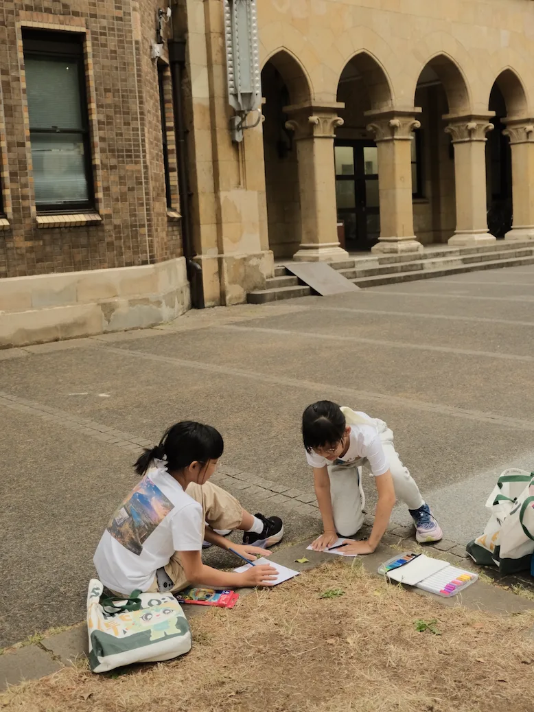 Two children kneeling on the pavement to sketch a red-brick arcaded campus building