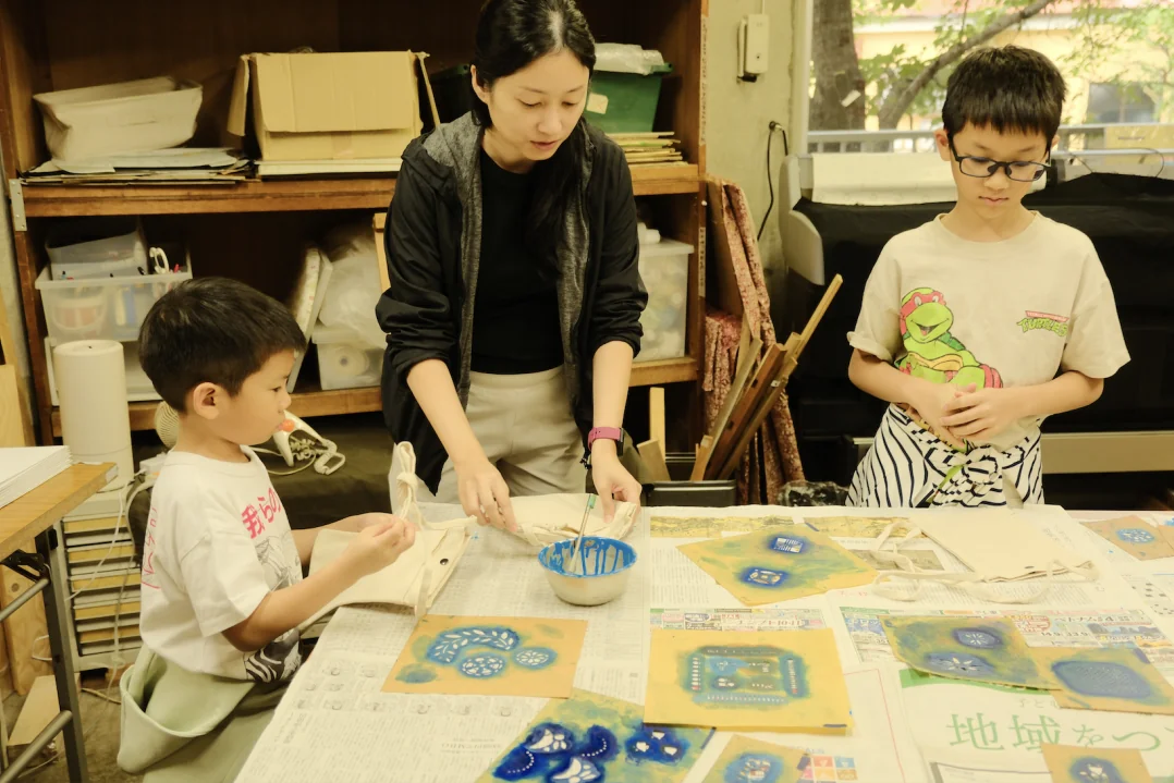 An instructor helping two boys dye tote bags on newspaper-covered worktables