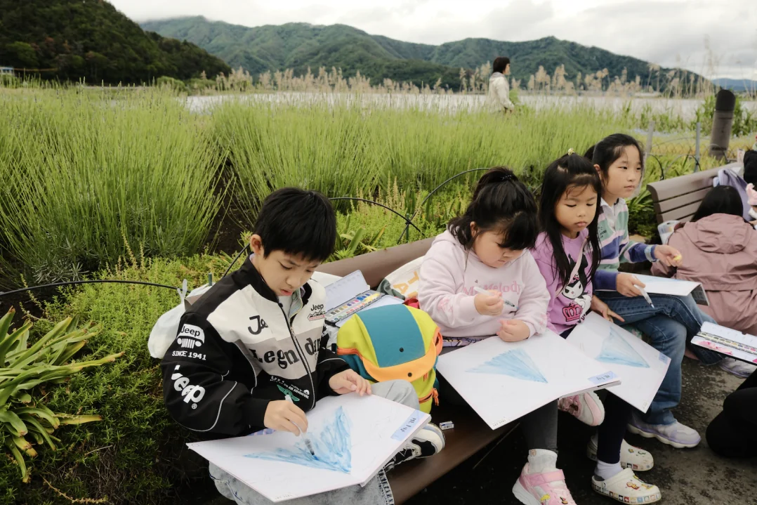 Children painting Mount Fuji on a lakeside bench with hills and pampas grass behind