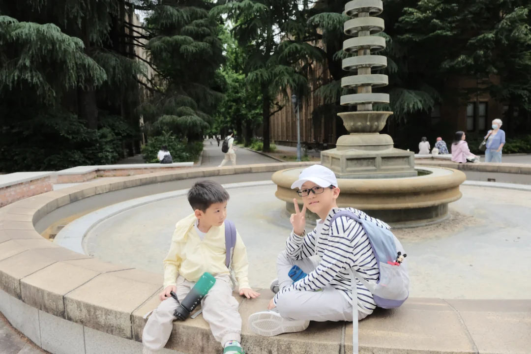 Two boys sitting on the rim of a stone fountain on the tree-lined campus
